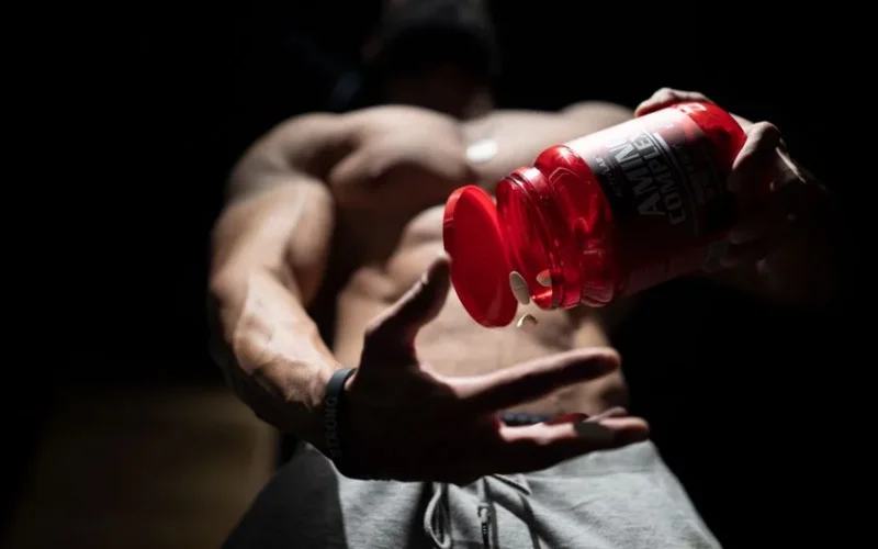 Shirtless muscular man pouring protein supplements from a bottle, showcasing fitness and strength.