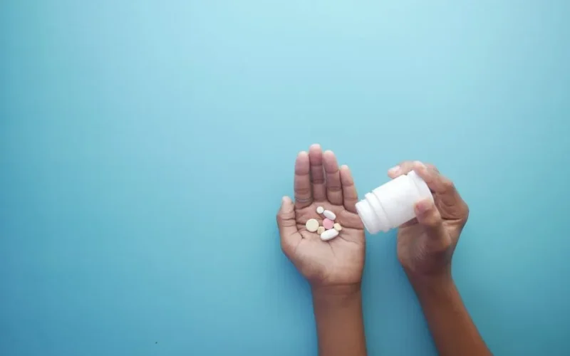Hands pouring pills from a bottle on a blue background, representing healthcare and medication.