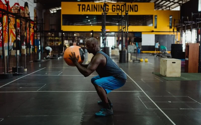 Side view of an African American man squatting with a medicine ball in an indoor gym setting, showcasing fitness and strength.