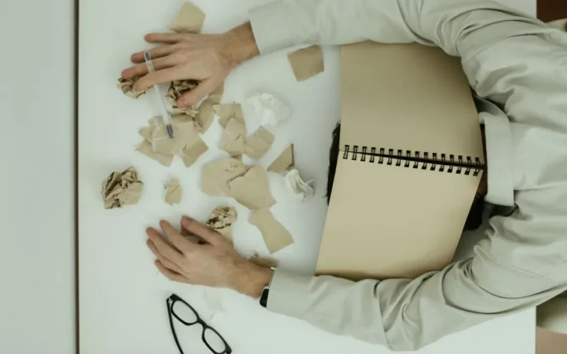 A tired adult man resting his head on a desk cluttered with crumpled papers and an open notebook.