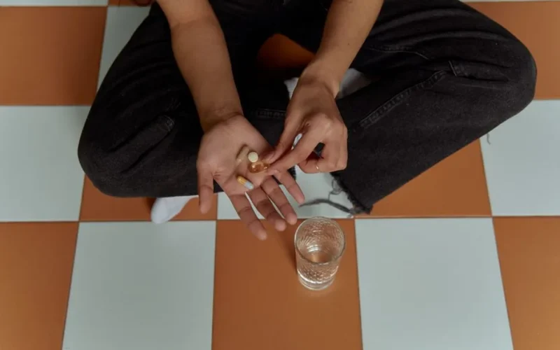 Individual sitting on checkered floor holding pills with a glass of water nearby.