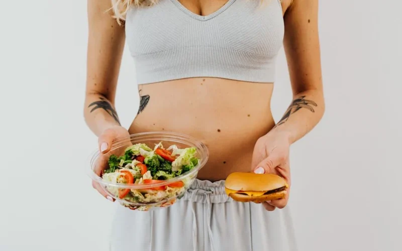 A woman in a sports bra holding a salad and cheeseburger, representing healthy vs junk food choices.