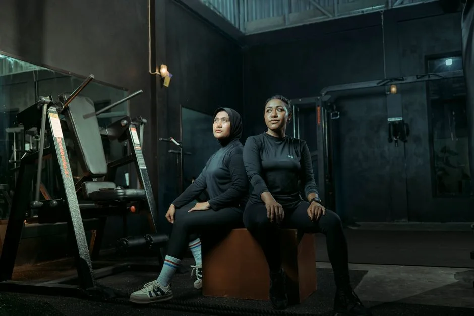 Two women in athletic wear sitting on a box with gym equipment in the background.