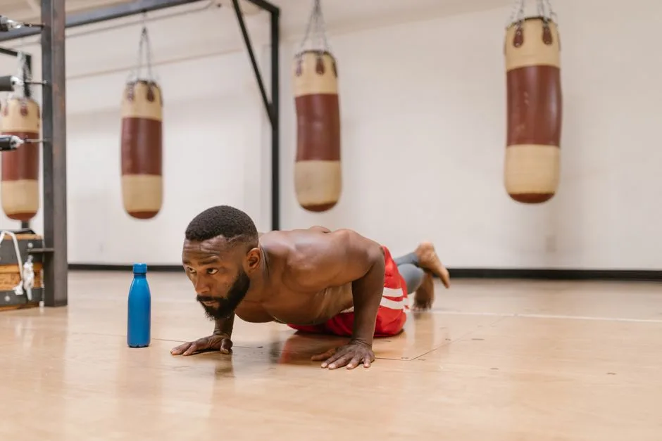 A focused shirtless man performing push-ups on a gym floor beside a blue water bottle.