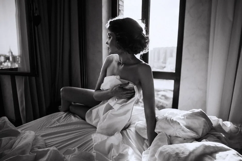 Moody black and white photo of a woman in a bedroom setting, captured with dramatic lighting.
