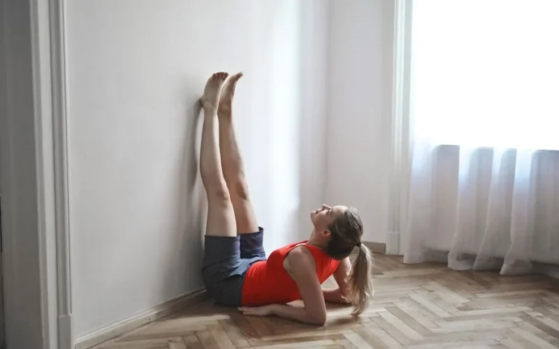 A woman in a red top performs a yoga stretch with legs vertical against a wall.