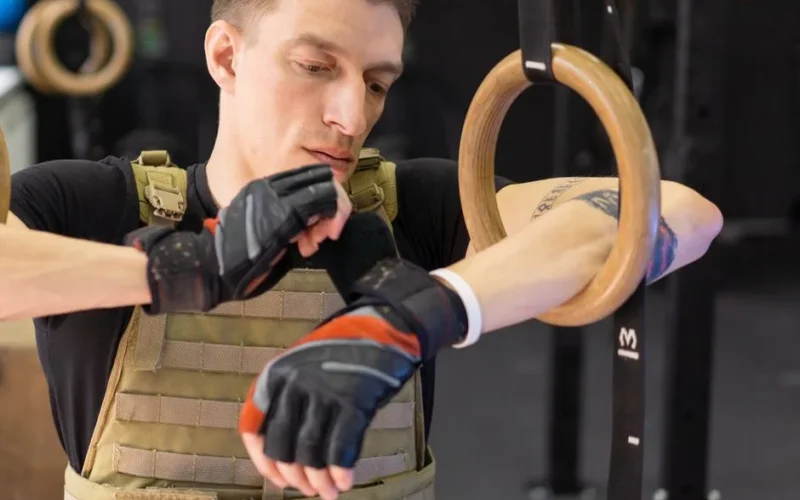 A man in tactical vest adjusting workout gloves next to gymnastic rings.