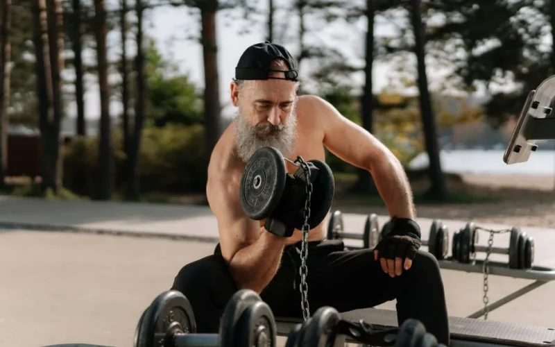 Elderly man lifting dumbbells in an outdoor gym setting, promoting healthy lifestyle.