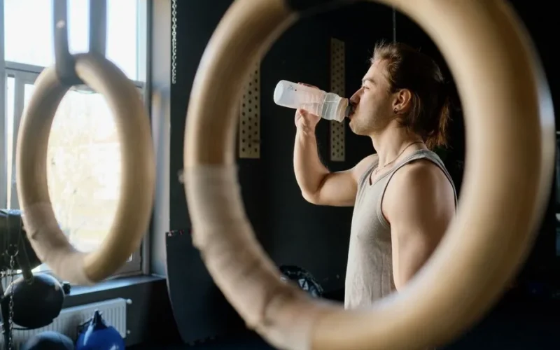 Athletic man drinking water in gym through gymnastic rings for hydration after workout.