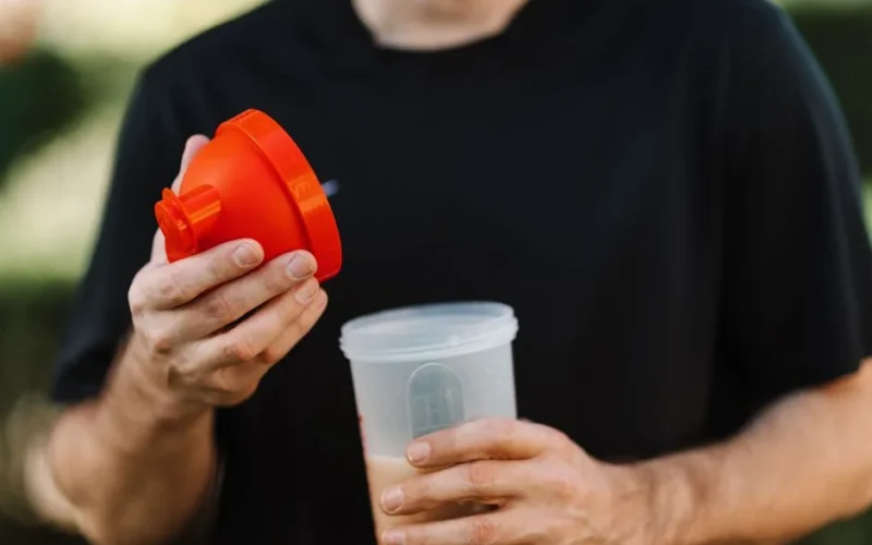 A man in a black shirt holds a protein shake tumbler with an orange lid, outdoors.