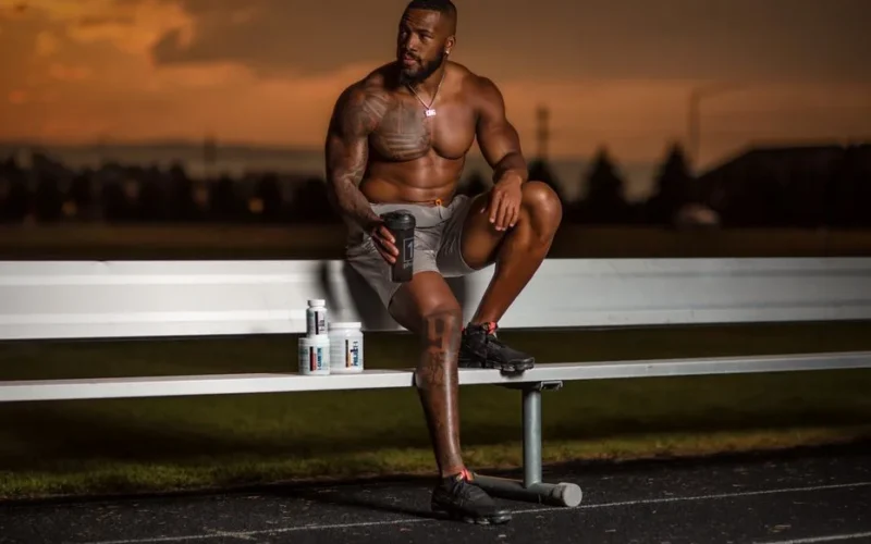 Muscular athlete sitting on a bench with supplements, enjoying a sunset at an outdoor stadium.