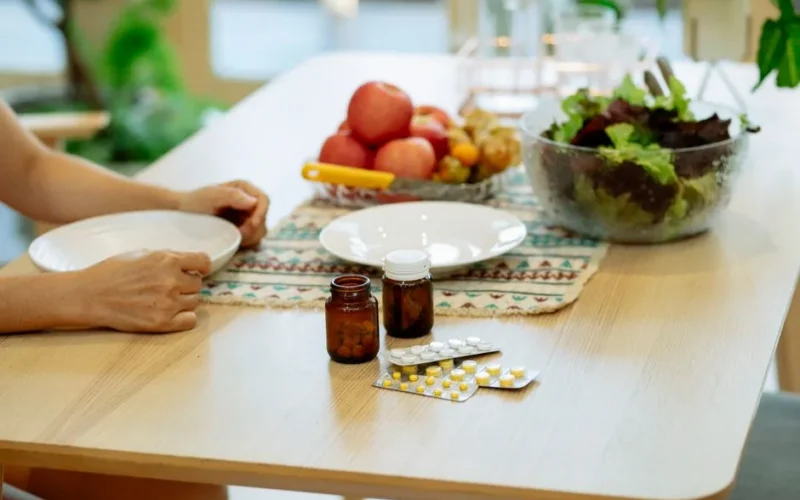 From above of crop anonymous person sitting at table with pile of various medicines and bowls of fresh lettuce salad and ripe fruits in kitchen