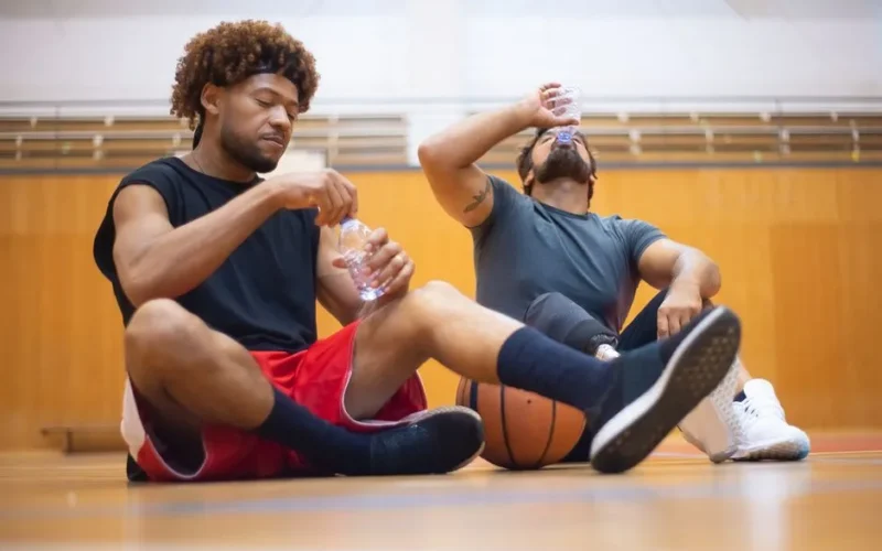 Two men sitting on indoor court, hydrating after playing basketball, showcasing sportsmanship.