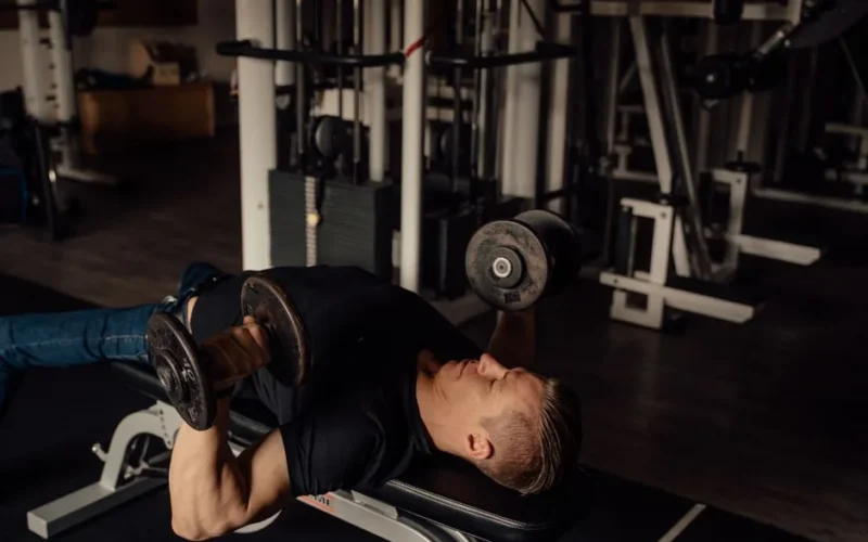 Muscular man doing dumbbell chest press exercise in a gym setting, highlighting fitness and strength.