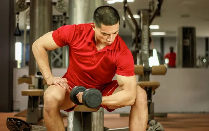 A muscular man in a red shirt lifts a dumbbell, focusing on bicep curls at an indoor gym.