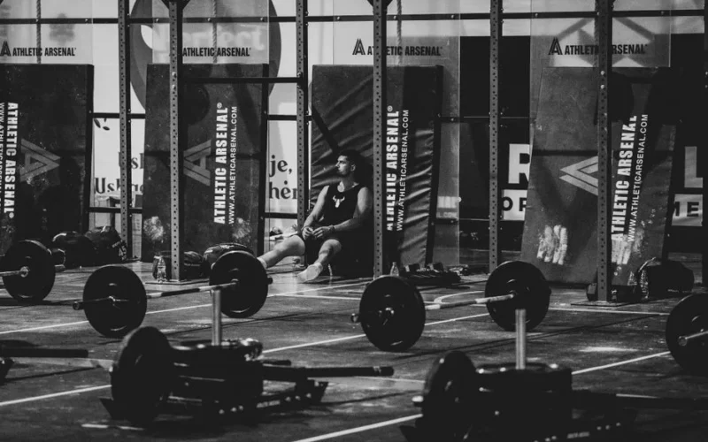 Black and white photo of a man resting in a gym surrounded by weights.