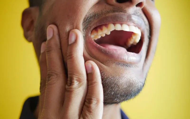 Close-up of a man holding his cheek, displaying a wide open mouth against a yellow background.