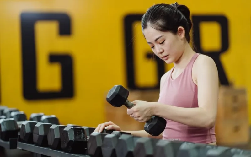Young woman lifting dumbbell in gym, focusing on strength training with sports equipment.