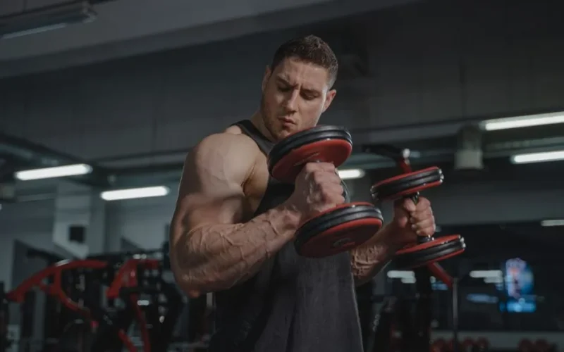 A strong man exercises with dumbbells, showcasing muscular build in a gym environment.