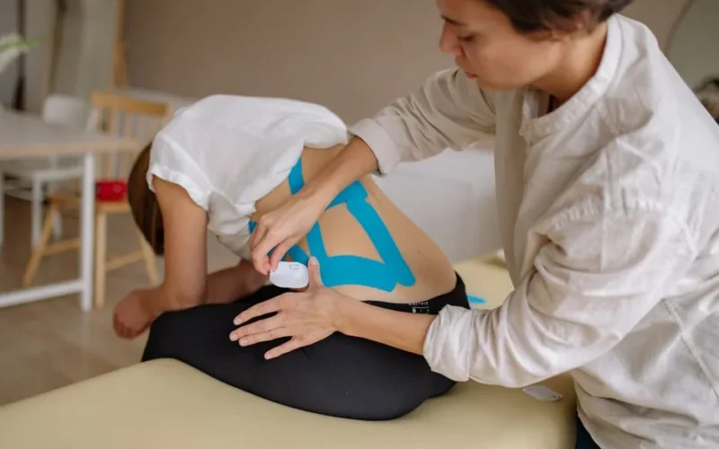 A therapist applies blue kinesiology tape to a patient's back in a clinical setting.