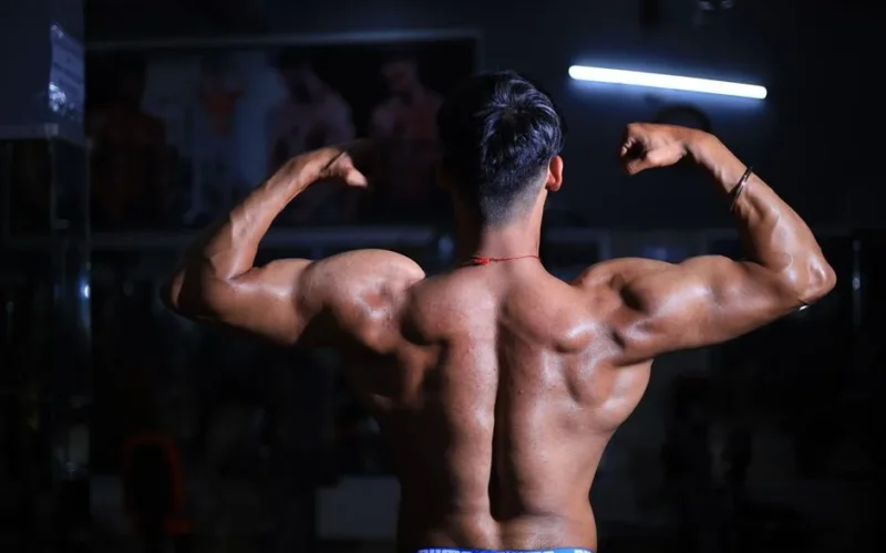 A muscular man flexes his back muscles in a dimly lit gym, showcasing strength and fitness.