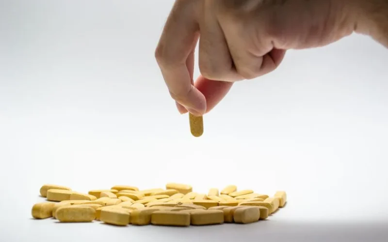 Close-up of a hand picking a yellow pill from a pile of tablets on a white surface.
