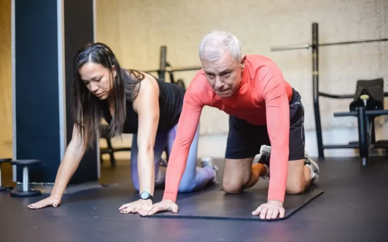Senior man and female trainer performing fitness exercises indoors, promoting healthy aging.