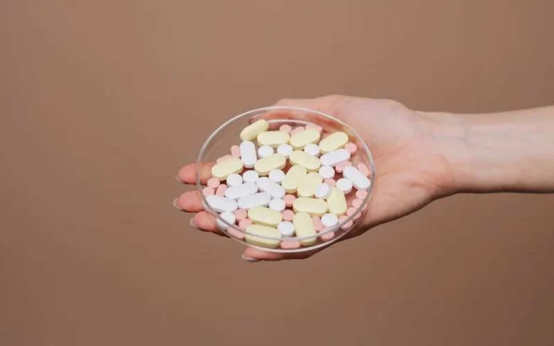 Close-up of a hand holding a petri dish filled with various tablets and capsules on a neutral background.