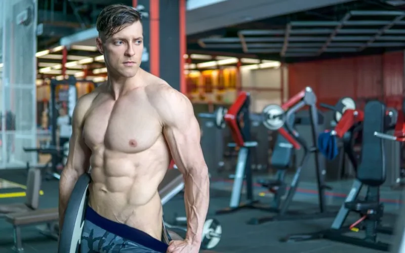 A muscular man poses in a gym showcasing his fit and toned physique, surrounded by exercise equipment.