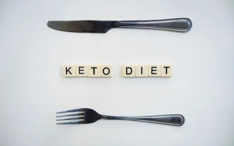 Top view of a knife, fork, and scrabble tiles spelling 'Keto Diet' on a white surface.