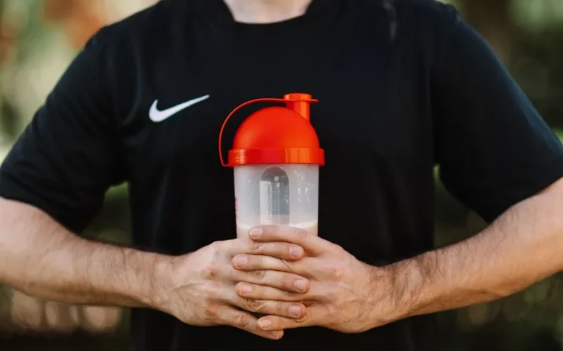 Close-up of a man holding a protein shaker outdoors, wearing a black shirt.