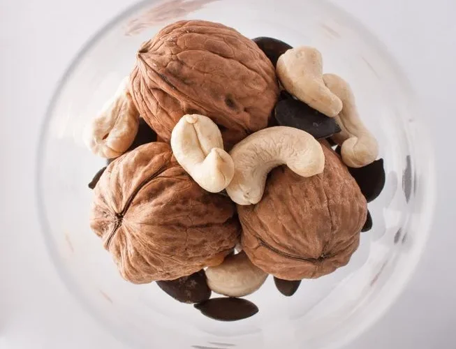 A close-up top view of assorted nuts including walnuts, cashews, and pumpkin seeds in a glass bowl.