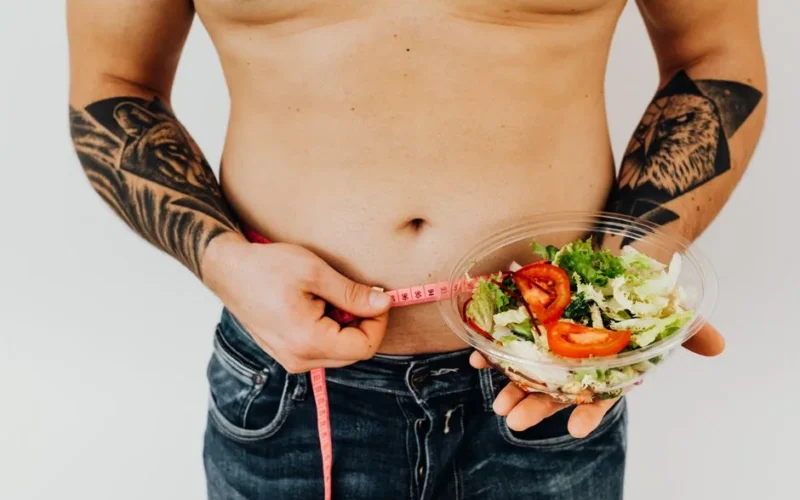 Tattooed man measuring waist while holding fresh salad, symbolizing healthy lifestyle and weight management.