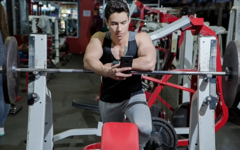 A young man resting in a gym while using a mobile phone beside weightlifting equipment.