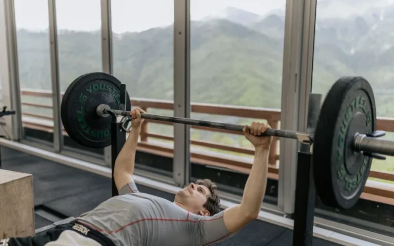 A man performs a bench press in a gym with mountain views, emphasizing strength and fitness.