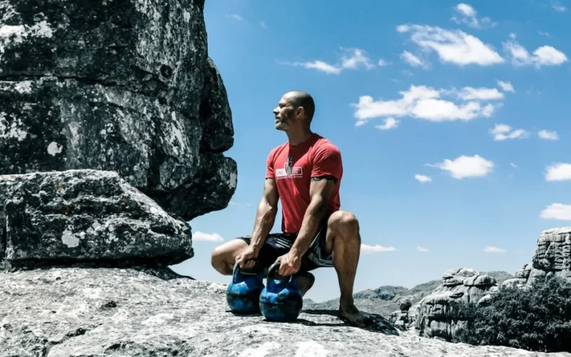 A muscular man exercises with kettlebells on rocky ground under a bright blue sky, emphasizing strength and fitness.
