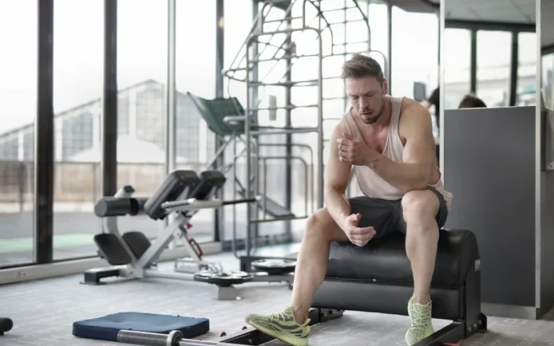 Tired young muscular male athlete in casual sportswear and sneakers sitting on bench and resting after intense weightlifting workout in modern fitness club