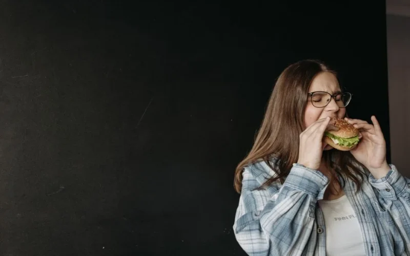 Woman in casual attire eating a hamburger in front of a dark background.
