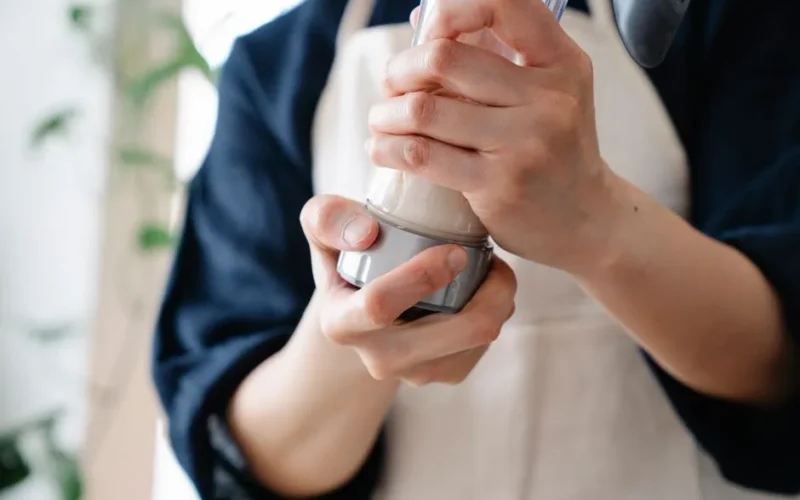 A person wearing an apron holds and shakes a protein shaker indoors. Casual food preparation scene.