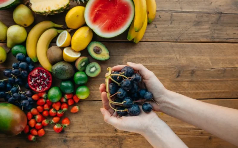 A variety of fresh, organic fruits with hands holding grapes on a wooden table.