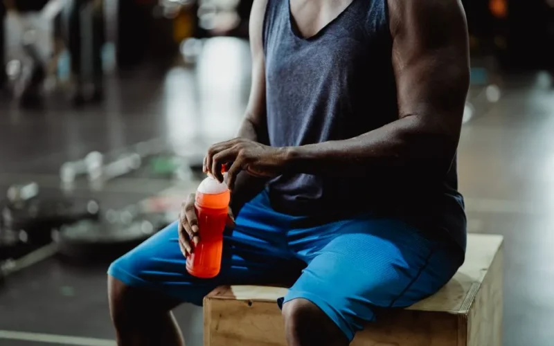 Muscular man taking a break with a sports drink in a gym setting.