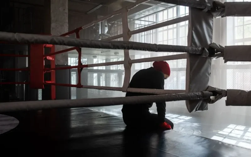 A boxer sitting alone in an empty gym boxing ring, enveloped in sunlight.