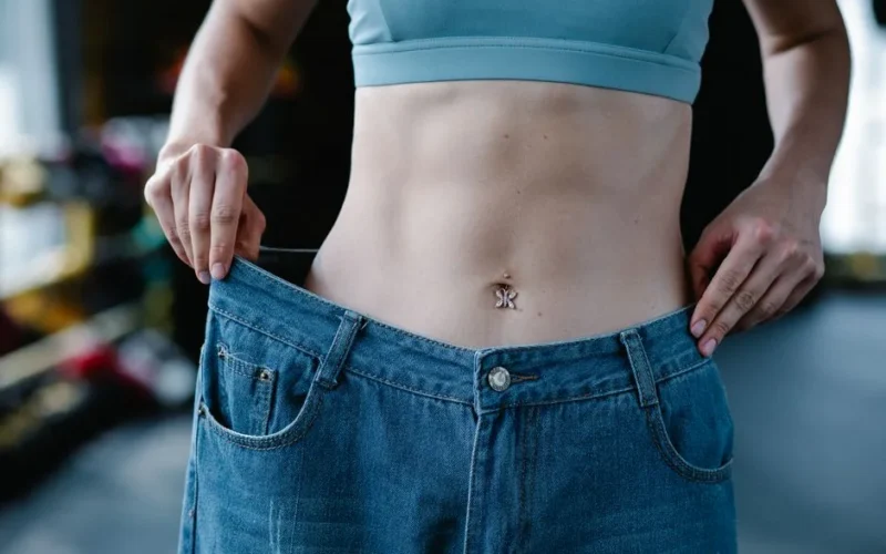 A woman shows her weight loss by holding oversized jeans revealing her toned stomach.