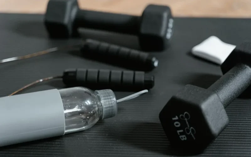 Close-up of gym essentials: dumbbells, jump rope, and water bottle on a mat.