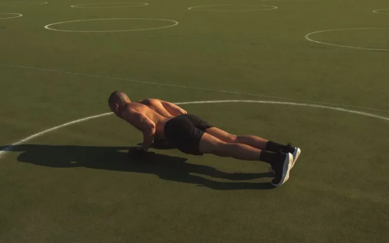 A shirtless man performing push-ups on a green field under bright daylight.