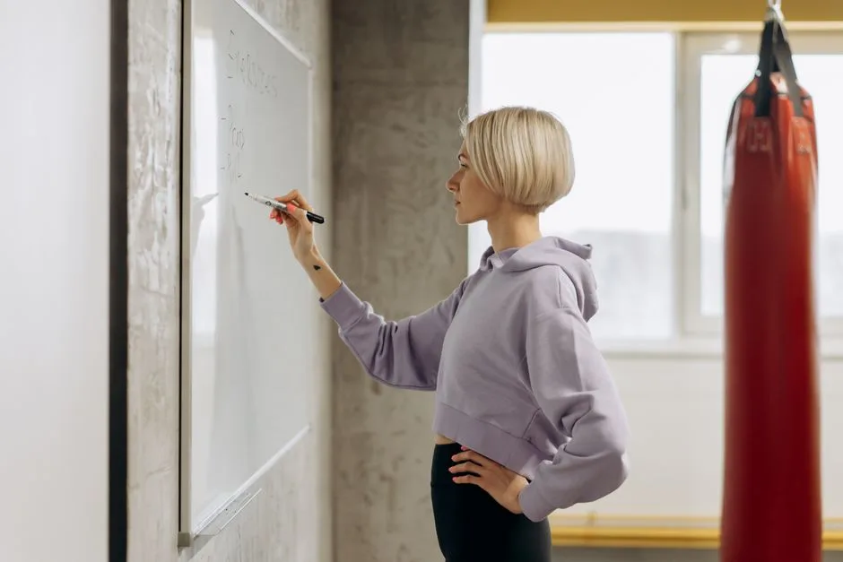 Fit female trainer writes workout plan on a whiteboard in a gym setting. Side view.